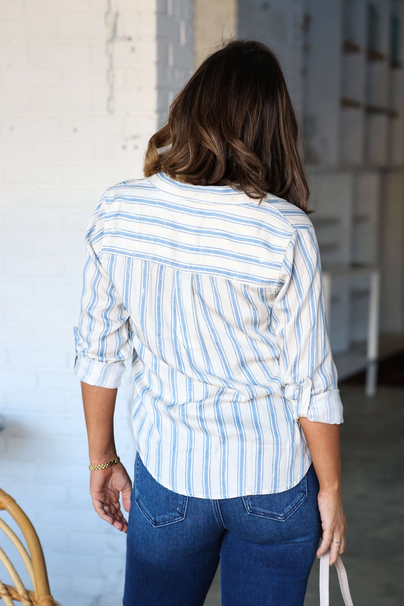 A woman with wavy brown hair wears the Thread and Supply Anna Blue Striped Shirt and blue jeans, seen from behind indoors.