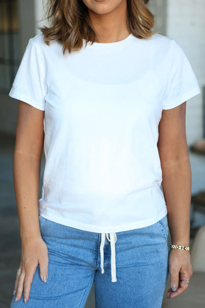 A woman in a Thread and Supply Basic White Tee with a round neckline and blue jeans stands indoors, her left hand by her side.