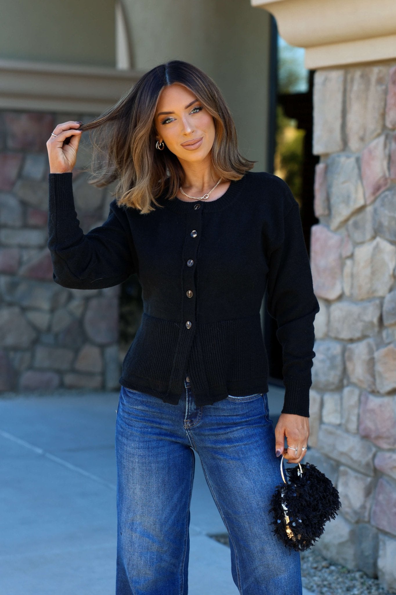 Woman with shoulder-length brown hair wears the Thread and Supply Black Button Down Cardigan and blue jeans by a stone wall, holding a black bag.