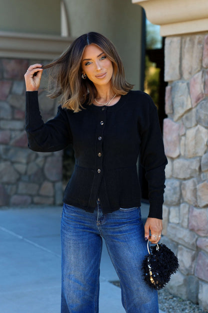 Woman with shoulder-length brown hair wears the Thread and Supply Black Button Down Cardigan and blue jeans by a stone wall, holding a black bag.