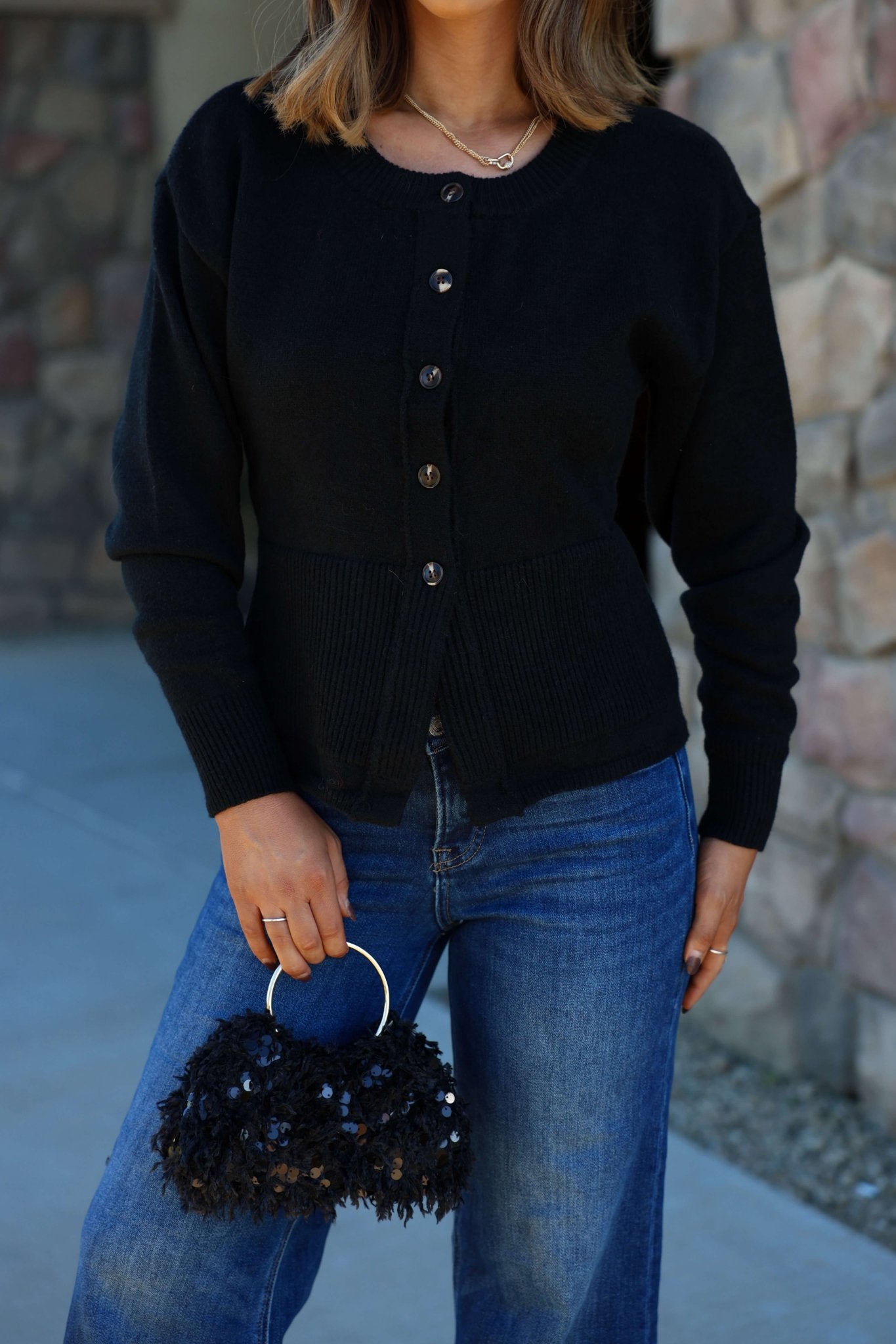 A woman in a Thread and Supply Black Button Down Cardigan and blue jeans holds a textured handbag by a stone wall outdoors.