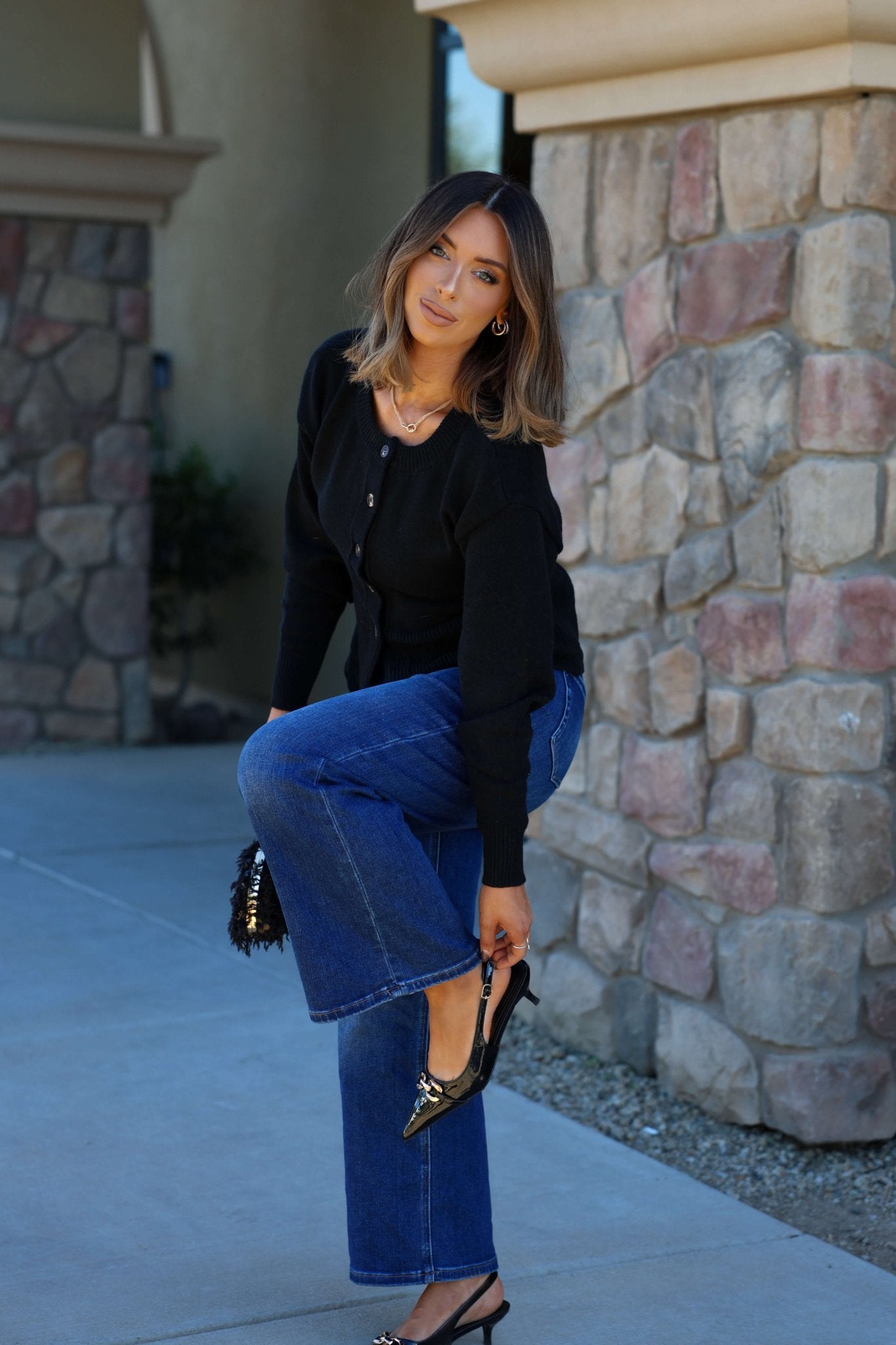 A woman wears the Thread and Supply Black Button Down Cardigan with blue jeans as she adjusts her black shoe by a stone wall outside.