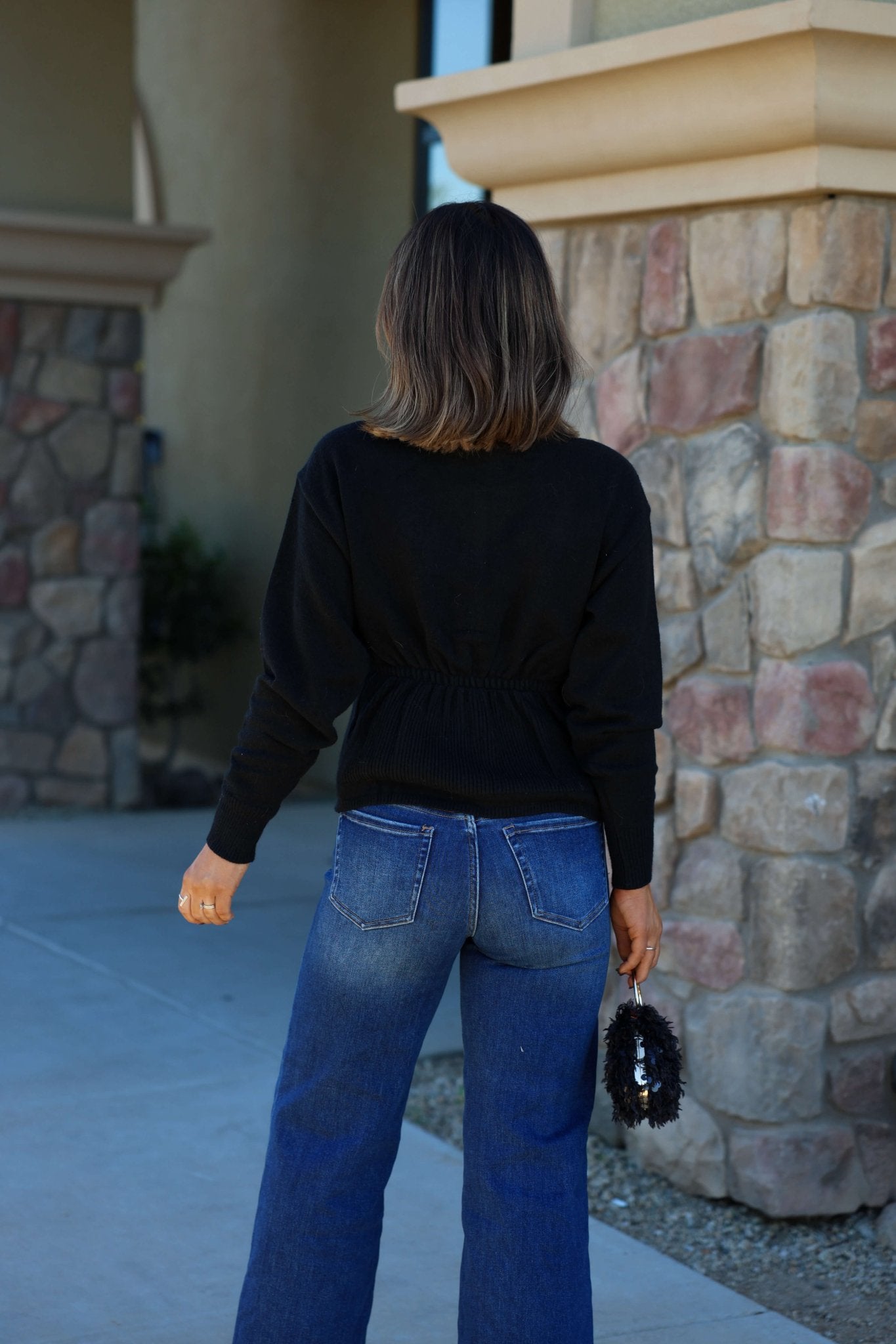 A woman in a Thread and Supply Black Button Down Cardigan and blue jeans stands outside, holding a small black accessory.