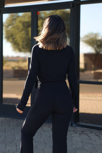 A woman with shoulder-length hair wears the Thread and Supply Black Cross Front Top outdoors, facing away by glass doors.