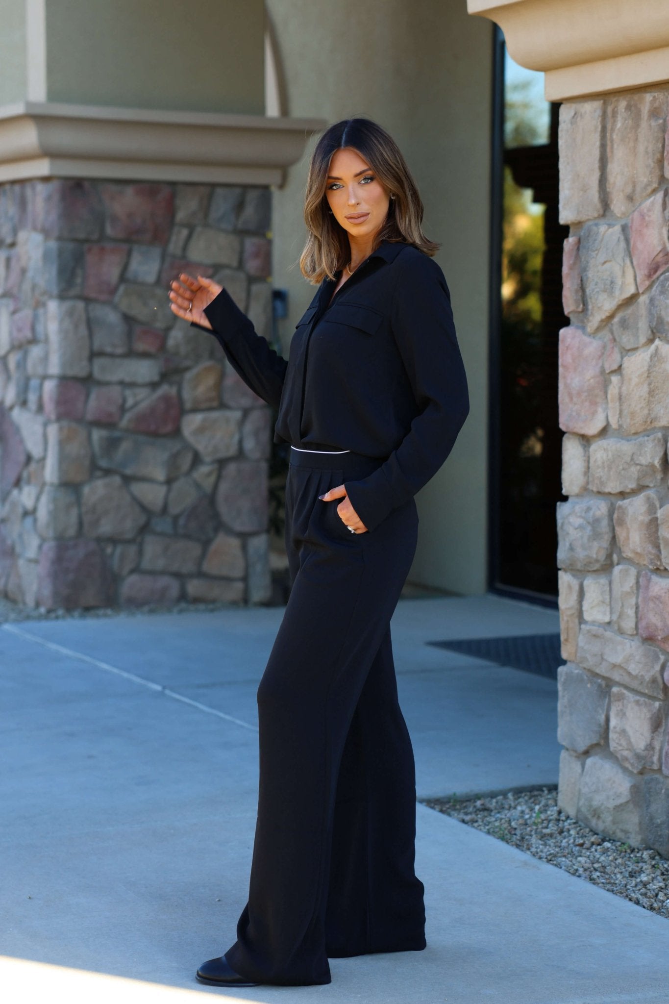Wearing Thread and Supply Black Dress Pants, a woman stands outdoors by a stone building, glancing back over her shoulder.