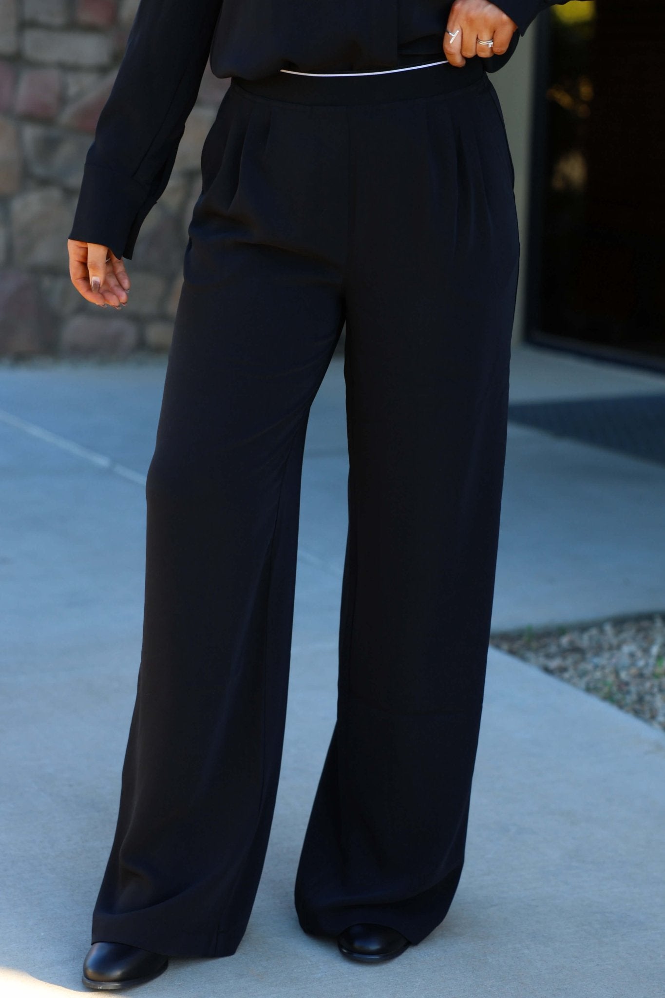 Someone wears Thread and Supply Black Dress Pants with an elastic waistband and black shoes, standing by a stone wall and glass door.