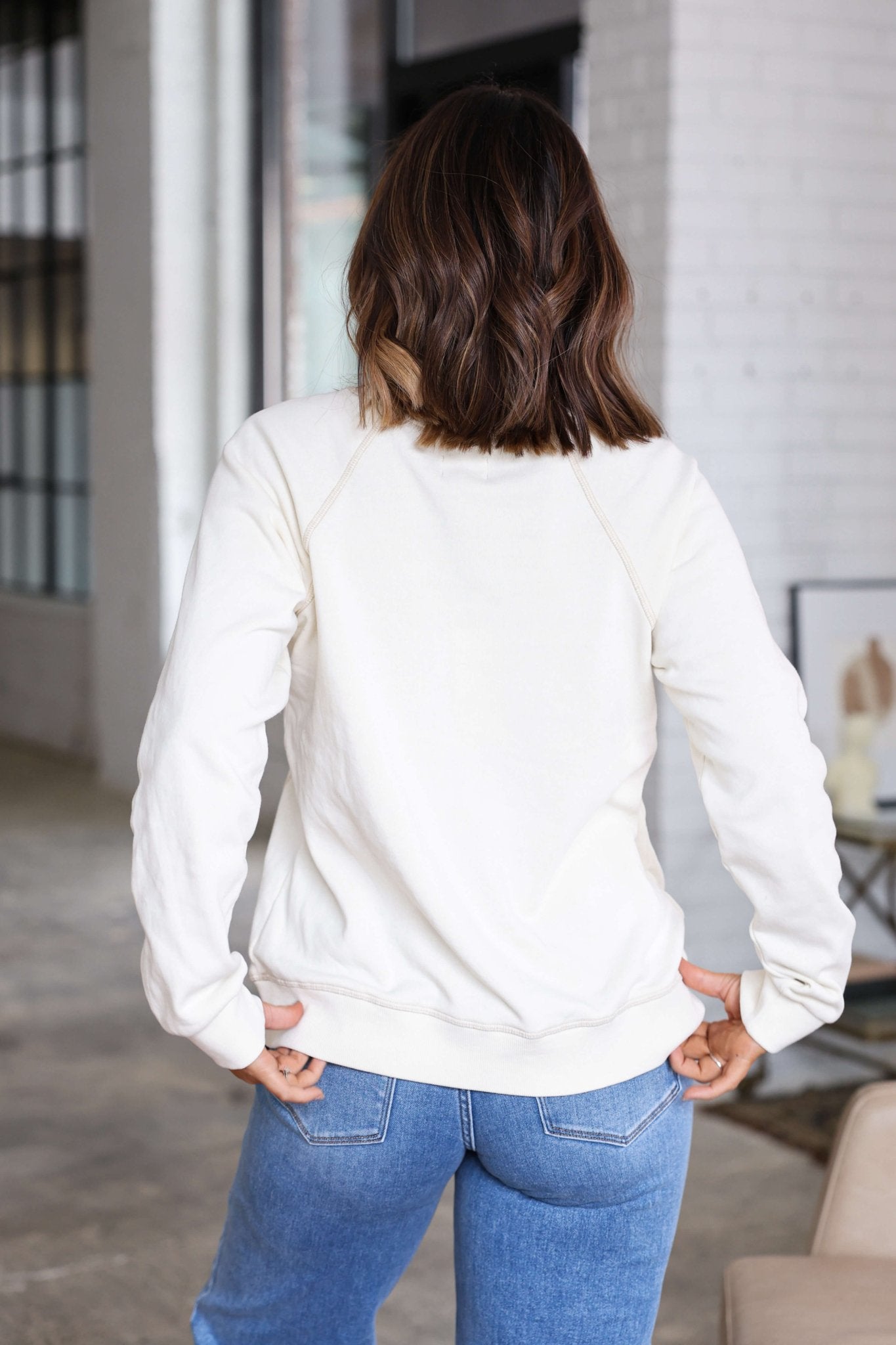 A woman with wavy brown hair wears a Thread and Supply Cream Ruby Sweatshirt and blue jeans, standing indoors, back to camera.