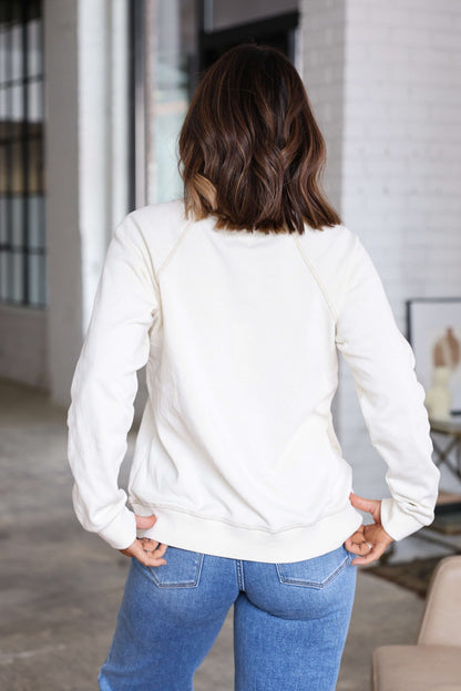A woman with wavy brown hair wears a Thread and Supply Cream Ruby Sweatshirt and blue jeans, standing indoors, back to camera.