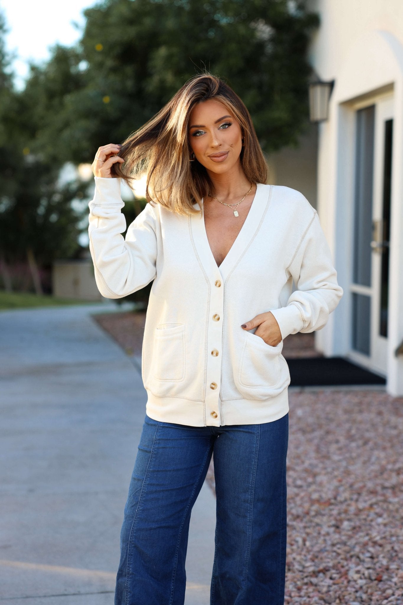 Woman with shoulder-length hair smiles outdoors, wearing the Thread and Supply Cream Stitch Buttoned Cardigan and blue jeans.