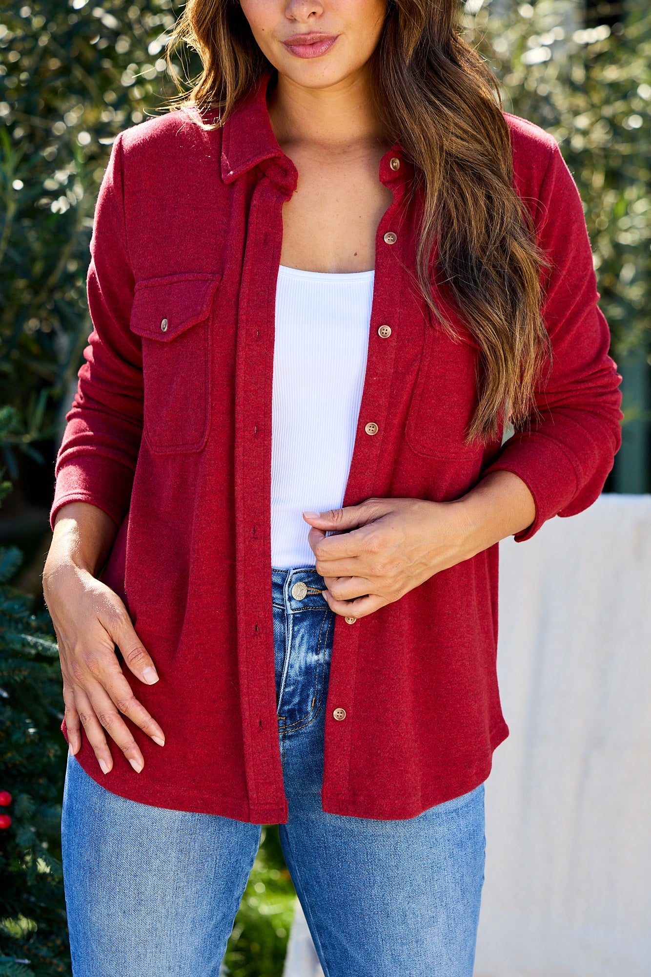 Woman wears the Thread and Supply Crimson Brushed Button Down Shirt over a white top and blue jeans, outdoors with greenery.