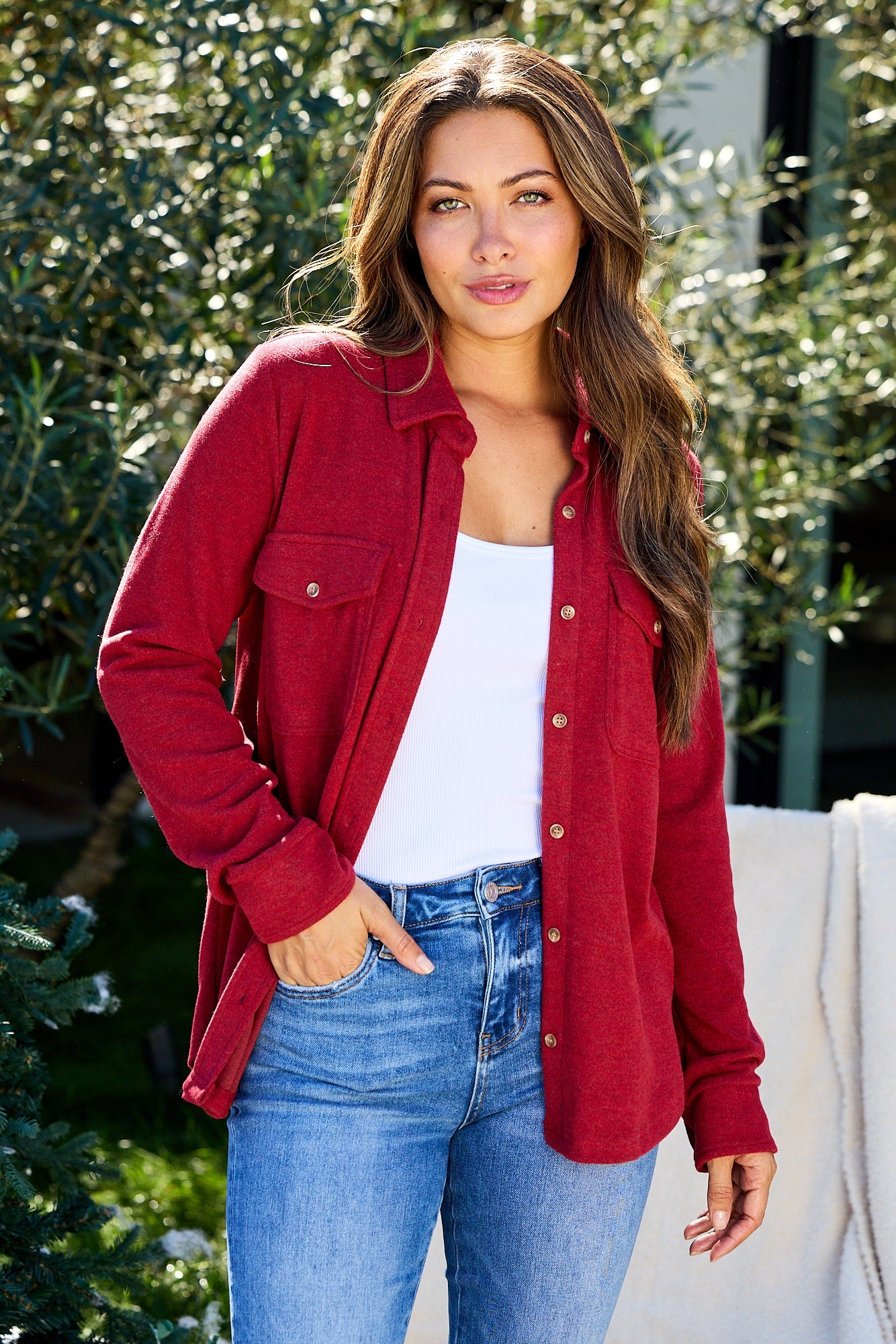 Woman with long brown hair in Thread and Supply Crimson Brushed Button Down Shirt, white top, and jeans stands outdoors among greenery.