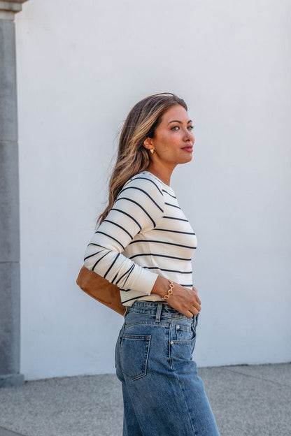 A woman in the Thread and Supply Ivory Striped Stacy Top and jeans stands sideways outdoors, showing off a casual style.