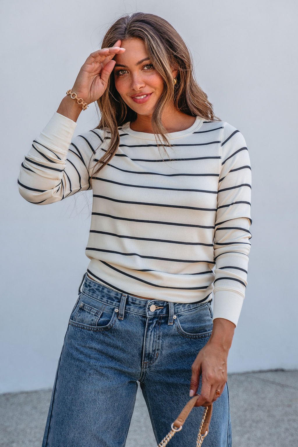 Woman in Thread and Supply Ivory Striped Stacy Top smiles in jeans, holding a purse and shielding her eyes in bright daylight.
