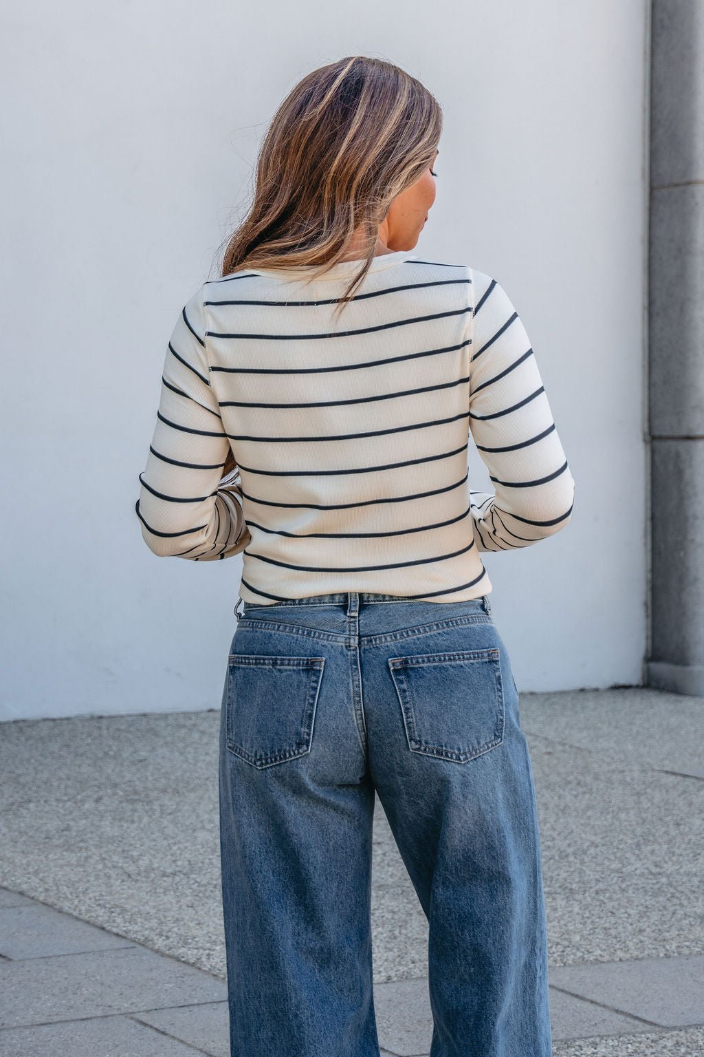 Someone with long hair, in blue jeans and a Thread and Supply Ivory Striped Stacy Top, stands outdoors facing away from the camera.