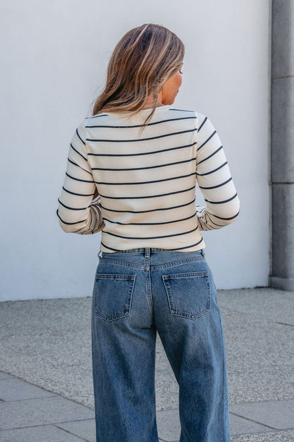 Someone with long hair, in blue jeans and a Thread and Supply Ivory Striped Stacy Top, stands outdoors facing away from the camera.
