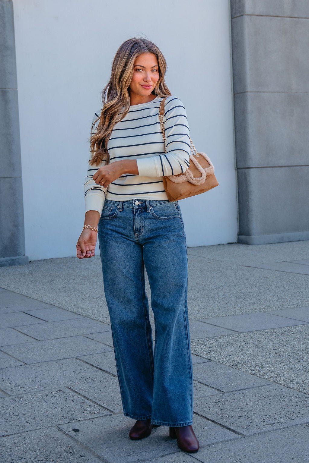 A woman in a Thread and Supply Ivory Striped Stacy Top and wide-leg jeans stands outside with brown boots and a tan handbag.