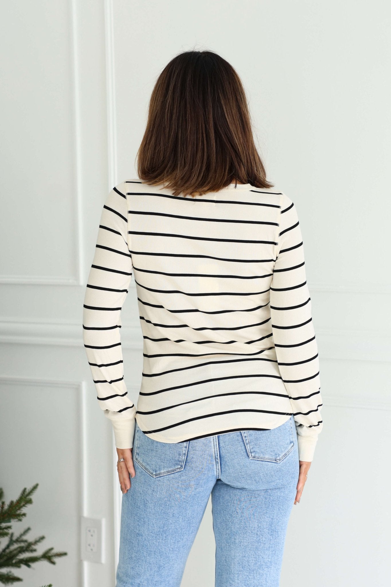 A woman faces away indoors wearing the Thread and Supply Ivory Striped Top with ribbed details and light blue jeans.