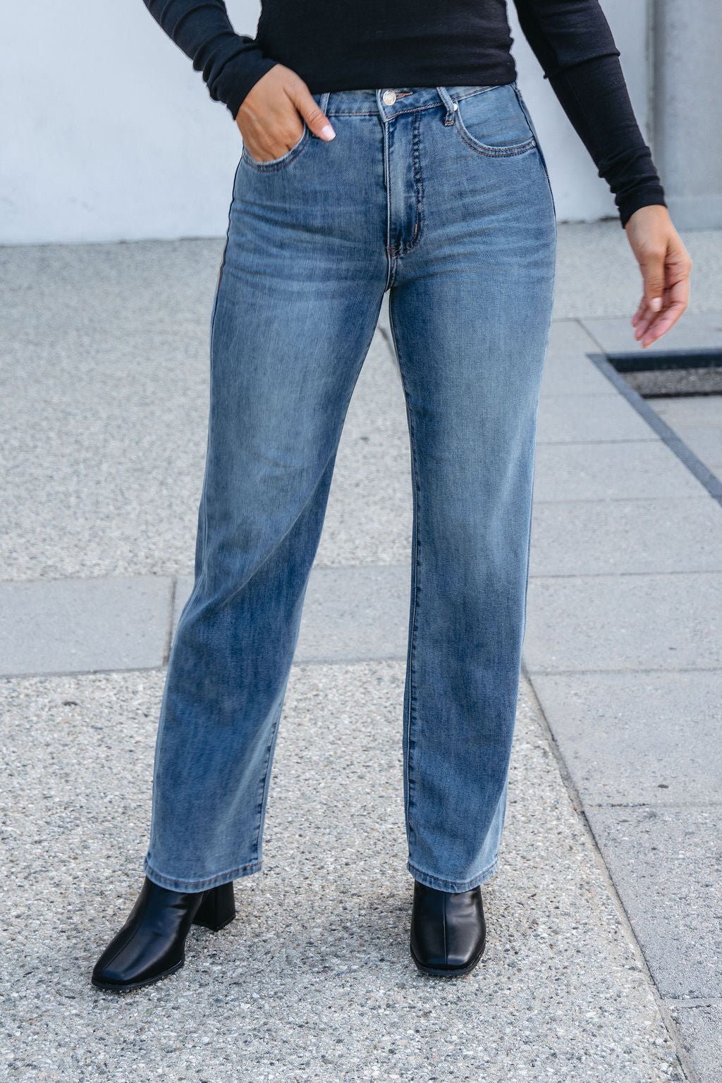Person wearing Thread and Supply Medium Wash Avery Straight Leg Jeans, black top, and heeled boots on a concrete outdoor surface.