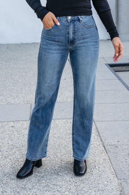 Person wearing Thread and Supply Medium Wash Avery Straight Leg Jeans, black top, and heeled boots on a concrete outdoor surface.