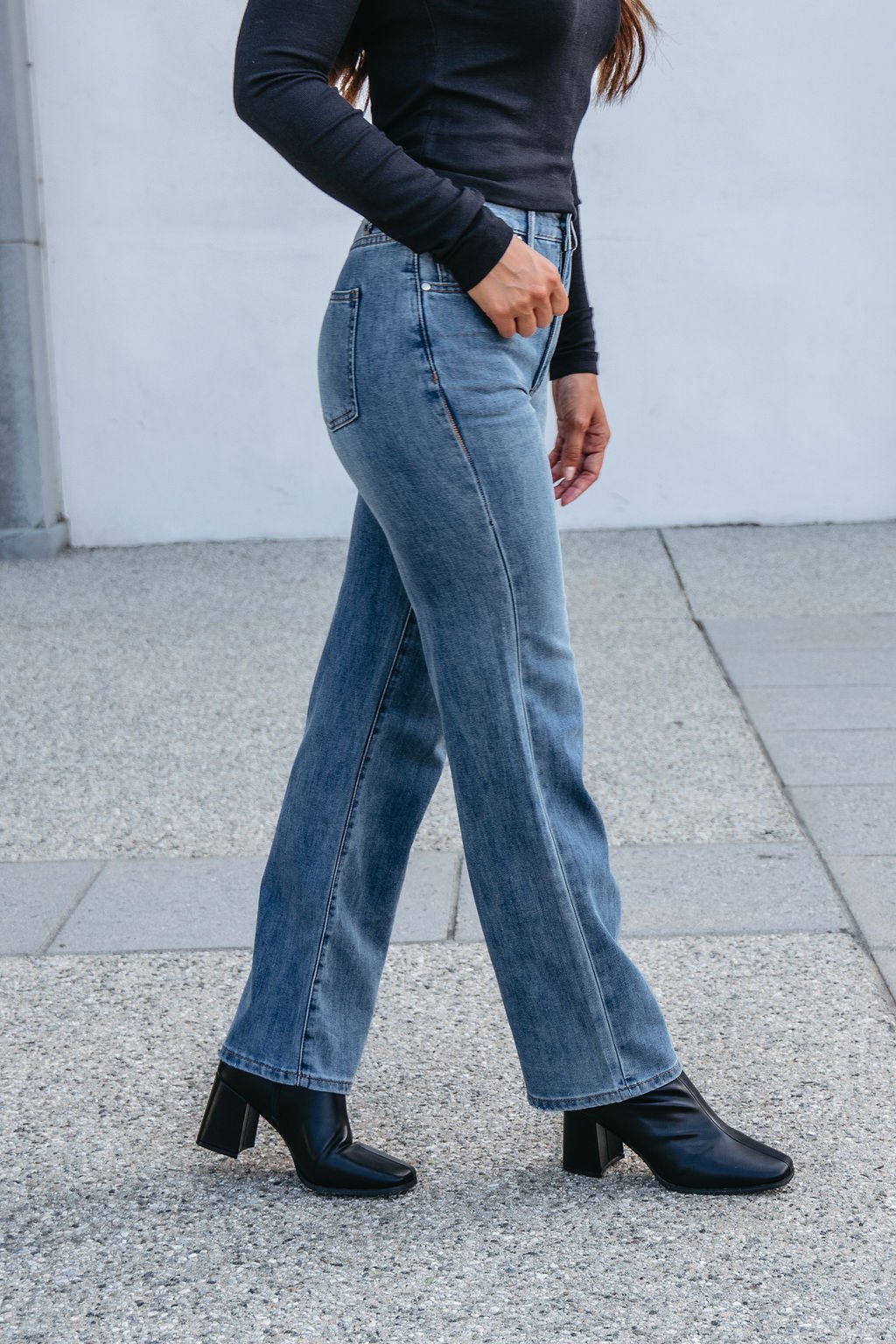 Person wearing Thread and Supply Medium Wash Avery Straight Leg Jeans, black long-sleeve top, and black heeled boots on a concrete sidewalk.