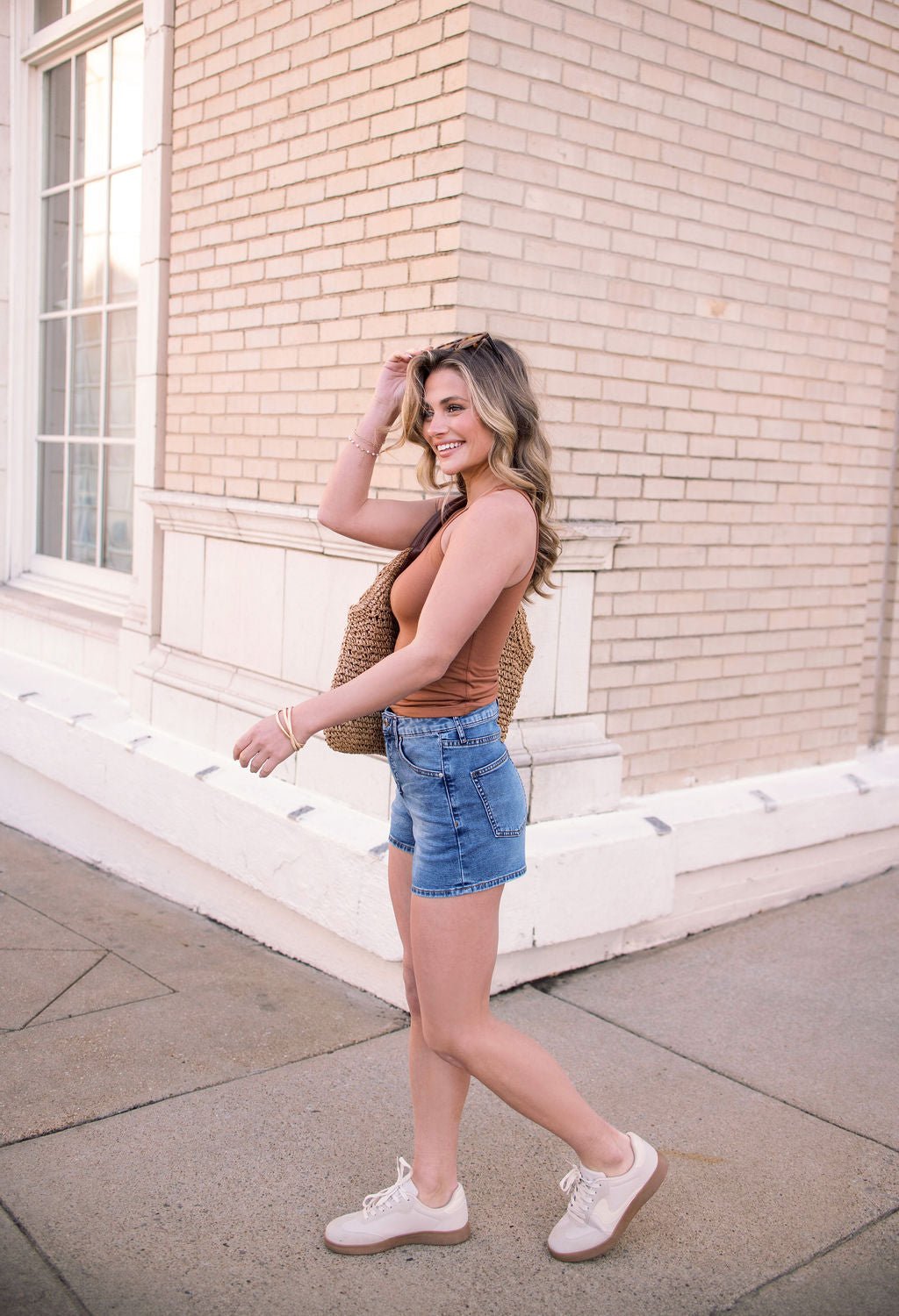 A smiling woman in a brown tank top, Thread and Supply Medium Wash Rowan Mid Rise Shorts, and sneakers walks outdoors with a woven bag.