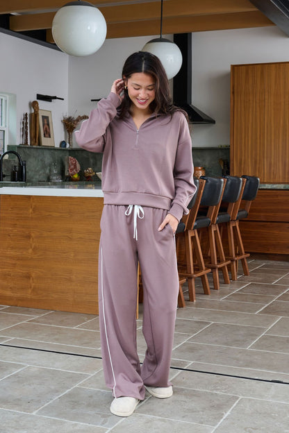 A woman in a Thread and Supply Mocha Hooded Quarter Zip Pullover smiles in a modern kitchen with wooden cabinets and bar stools.