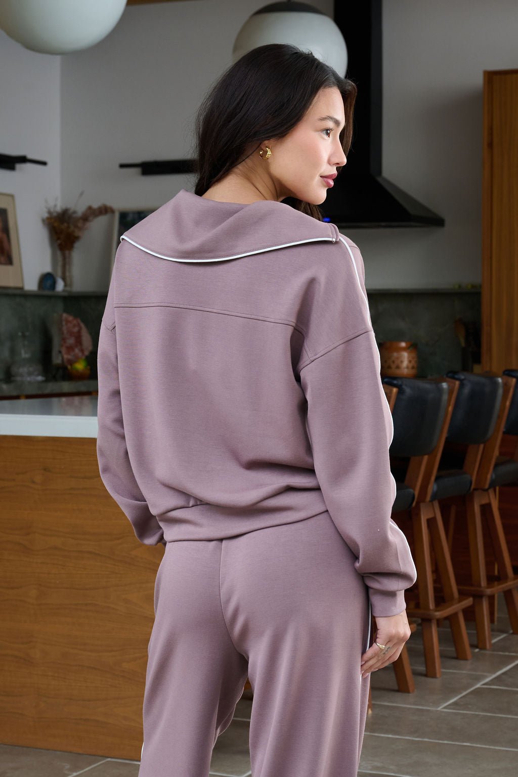 Woman in a Thread and Supply Mocha Hooded Quarter Zip Pullover stands in a modern kitchen, facing away with her head turned aside.