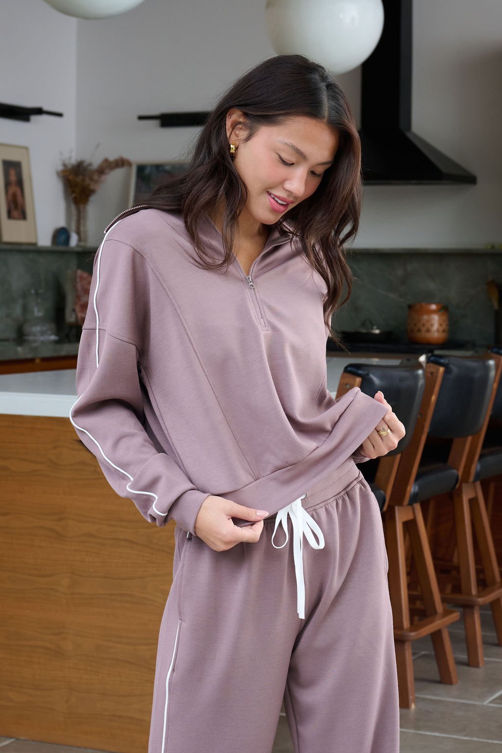 A woman in mauve loungewear smiles in a modern kitchen, wearing the Thread and Supply Mocha Hooded Quarter Zip Pullover.