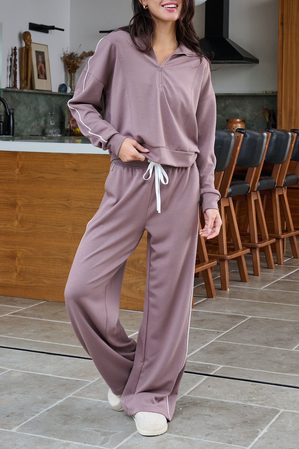 Woman wears Thread and Supply Mocha Wide Leg Drawstring Pants in a modern kitchen with wooden chairs and a green-tiled backsplash.