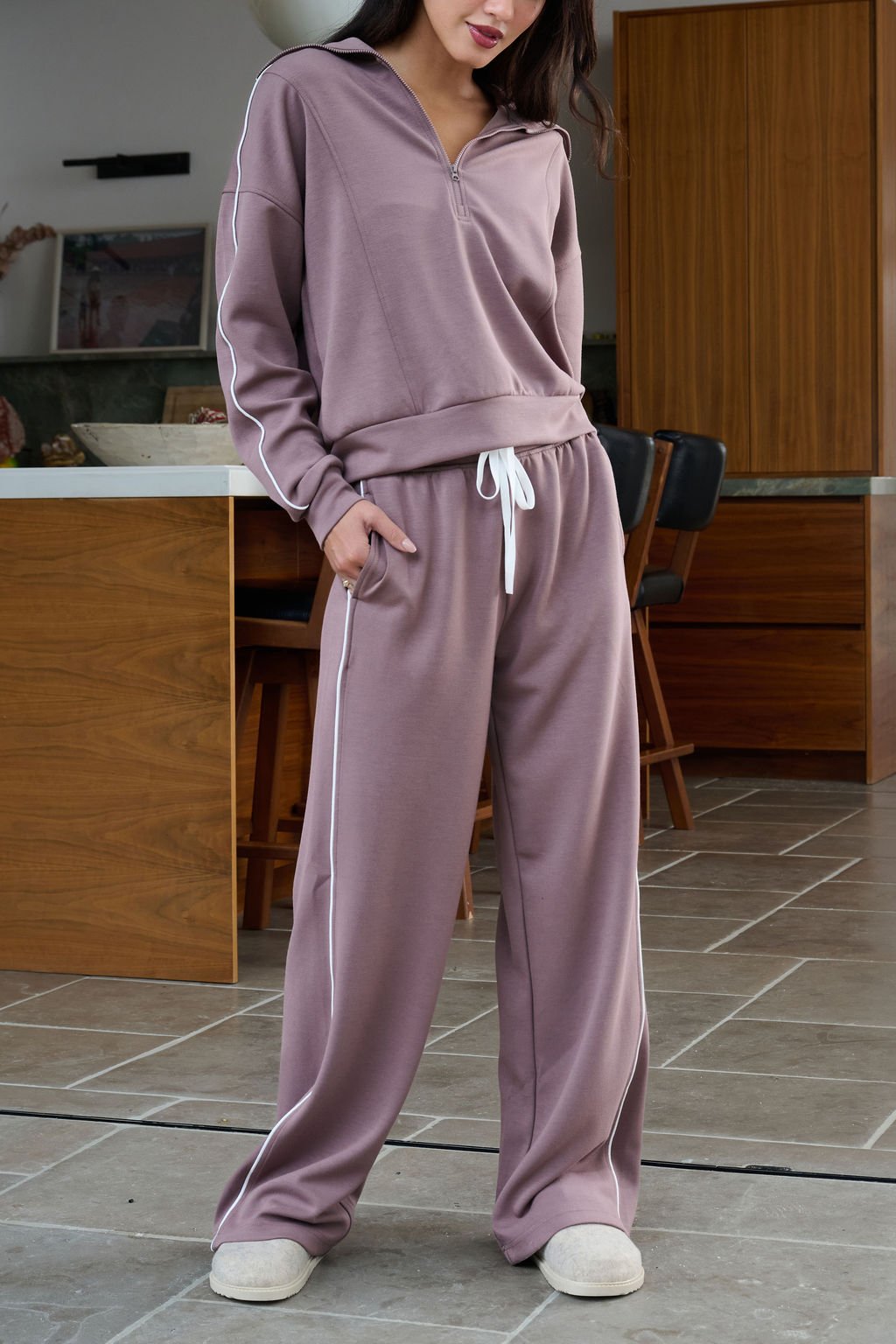 A woman in Thread and Supply Mocha Wide Leg Drawstring Pants stands on a tiled floor indoors, hands in her pockets.