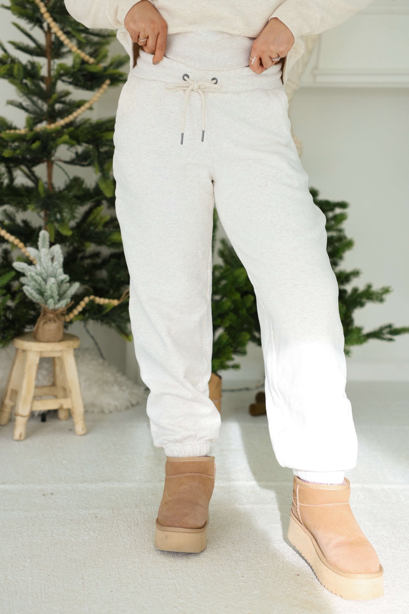 Person in Thread and Supply Oatmeal Fleece Joggers and tan boots stands indoors near green artificial Christmas trees, looking cozy.