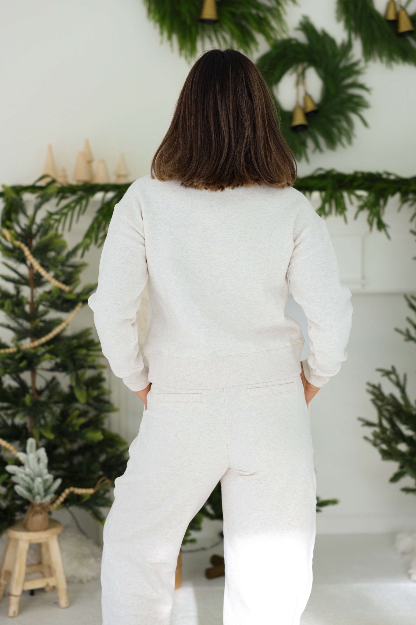 A person in a Thread and Supply Oatmeal Quarter Zip Sweatshirt stands with their back to green holiday decor and small trees.