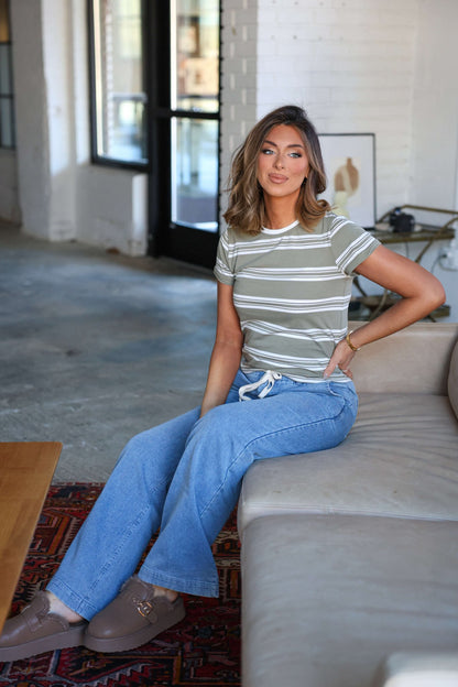 A woman in a Thread and Supply Olive Striped Short Sleeve Top and jeans sits on a beige couch in a bright, windowed room.