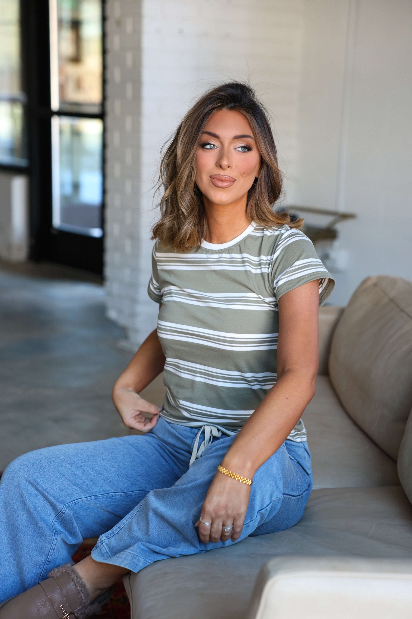Woman with wavy hair in Thread and Supply Olive Striped Short Sleeve Top and jeans smiles on a beige couch in a bright, modern room.