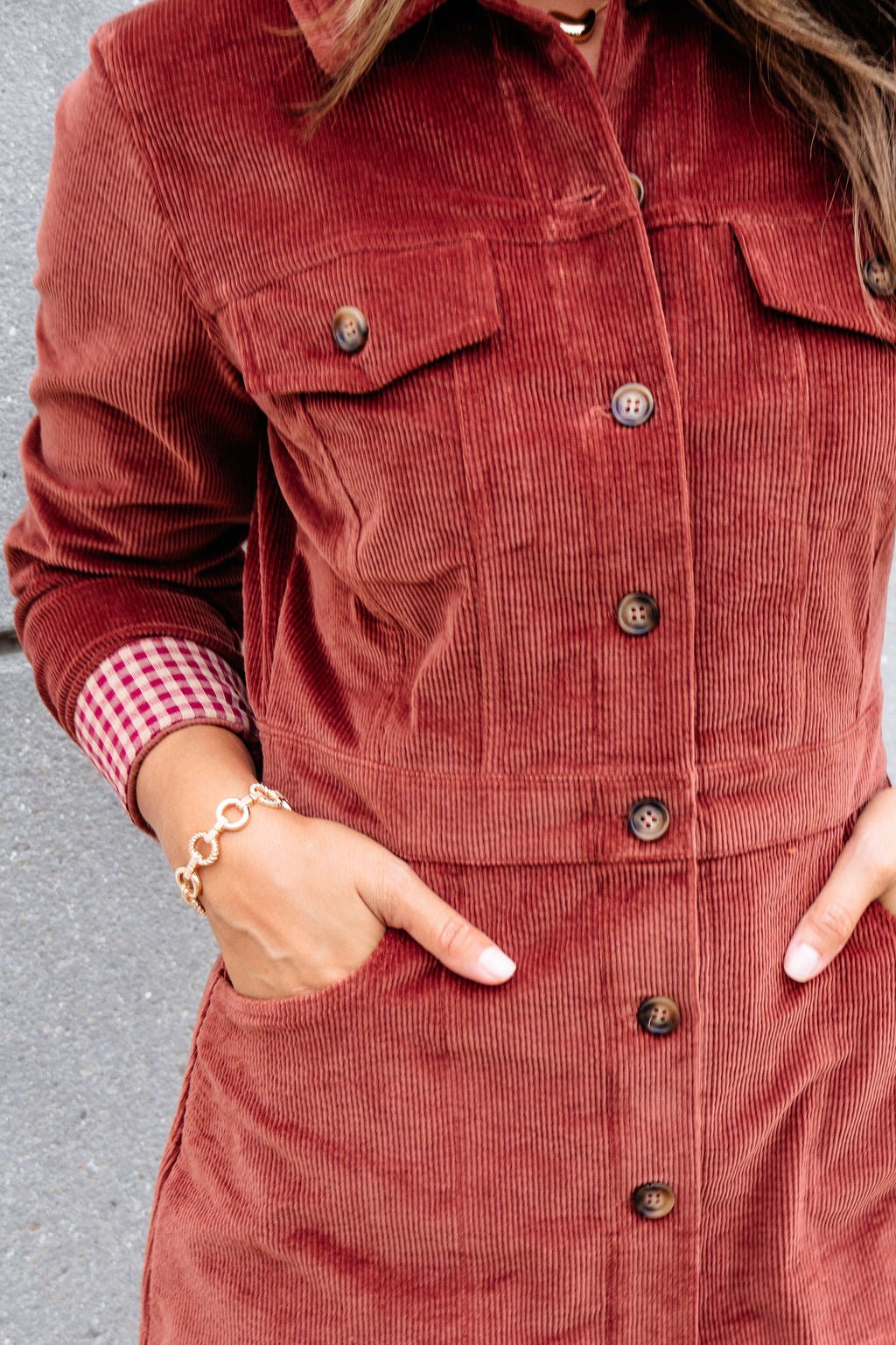 Wearing the Thread and Supply Rust Corduroy Mini Dress with front buttons, hands in pockets, checkered shirt cuff, and bracelet shown.