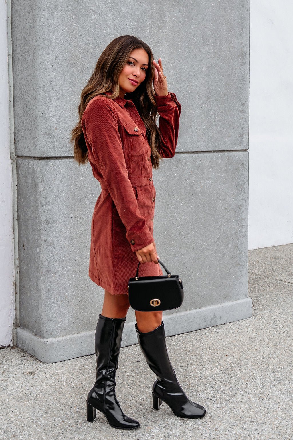 Woman in the Thread and Supply Rust Corduroy Mini Dress with knee-high black boots and a black handbag by a concrete wall.