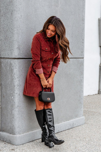 A woman smiles in the Thread and Supply Rust Corduroy Mini Dress, black knee-high boots, and holds a purse while leaning on a gray wall.