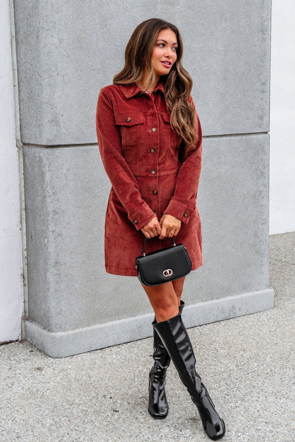 Woman in a Thread and Supply Rust Corduroy Mini Dress, black knee-high boots, and a black handbag stands by a concrete wall.
