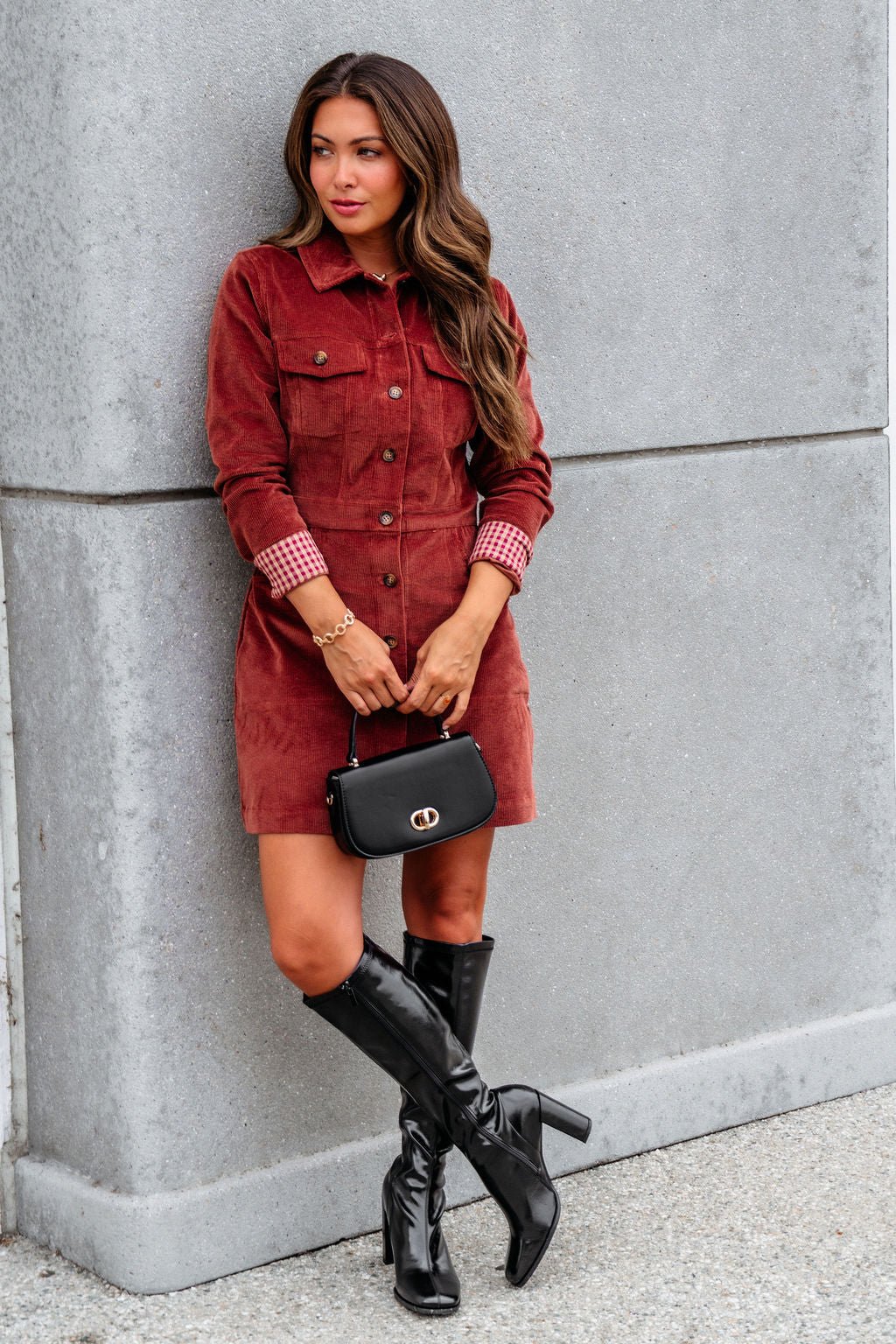 Woman in a Thread and Supply Rust Corduroy Mini Dress holds a black purse, wearing knee-high boots by a gray concrete wall.