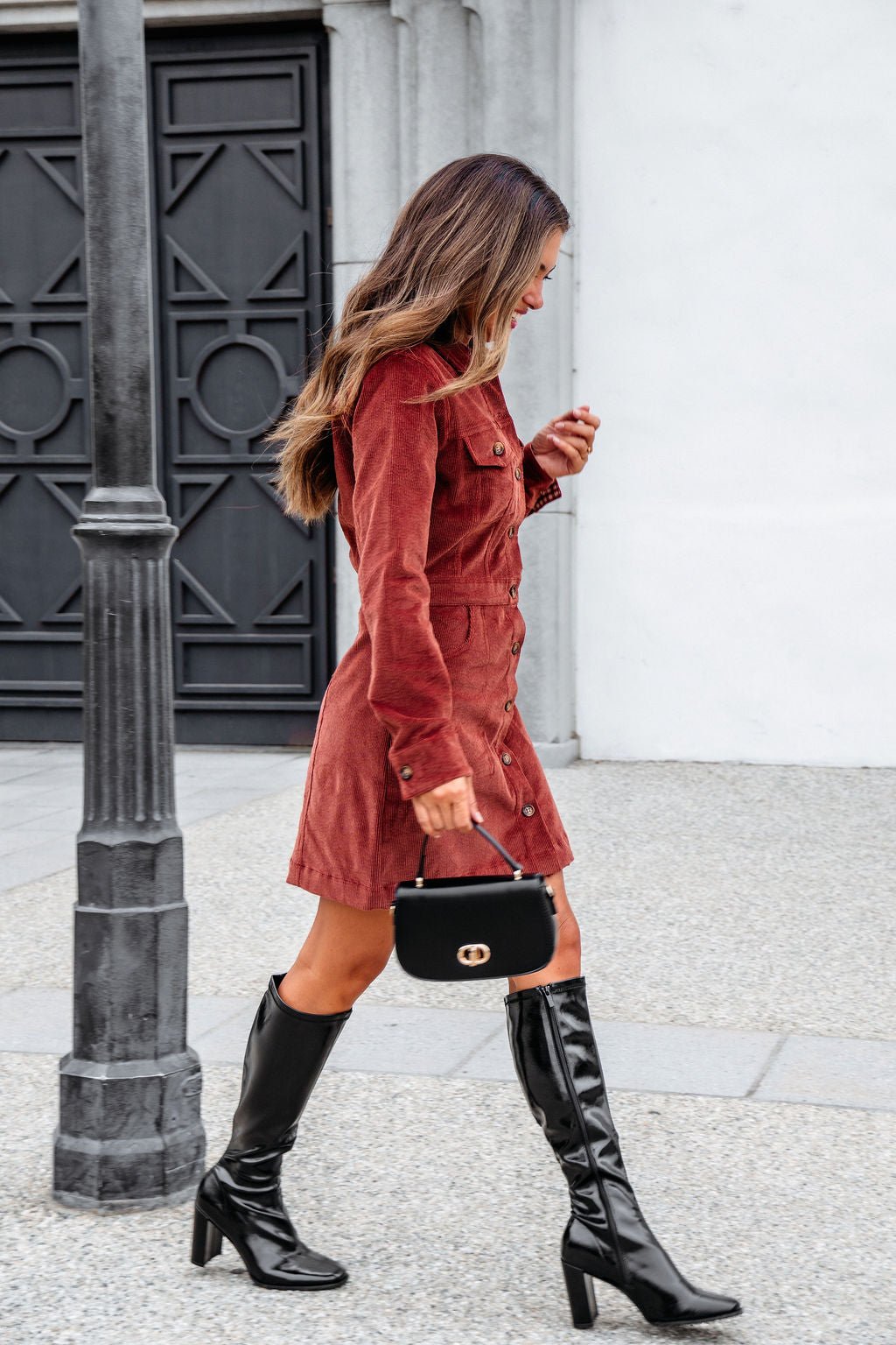 A woman in a Thread and Supply Rust Corduroy Mini Dress and black boots walks with a small black handbag past a lamp post.
