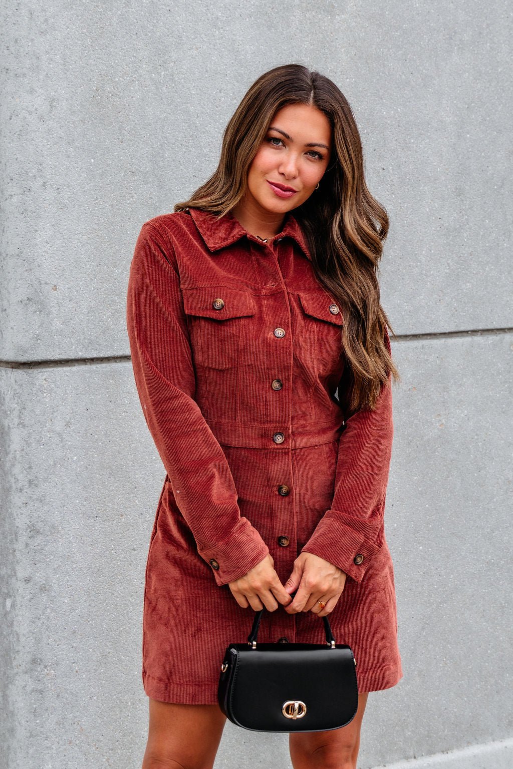 A woman in a Thread and Supply Rust Corduroy Mini Dress stands by a gray wall, holding a small black handbag.