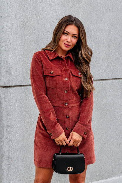 A woman in a Thread and Supply Rust Corduroy Mini Dress stands by a gray wall, holding a small black handbag.