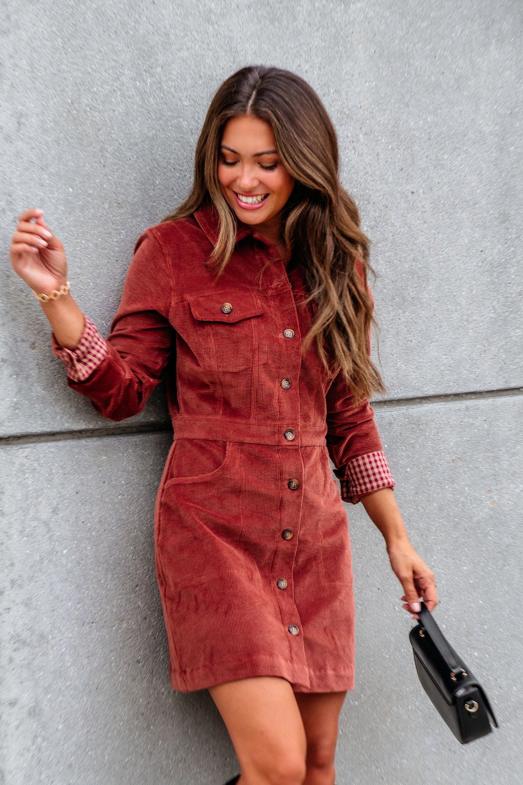 Woman wears the Thread and Supply Rust Corduroy Mini Dress, holding a black purse and smiling against a gray wall.