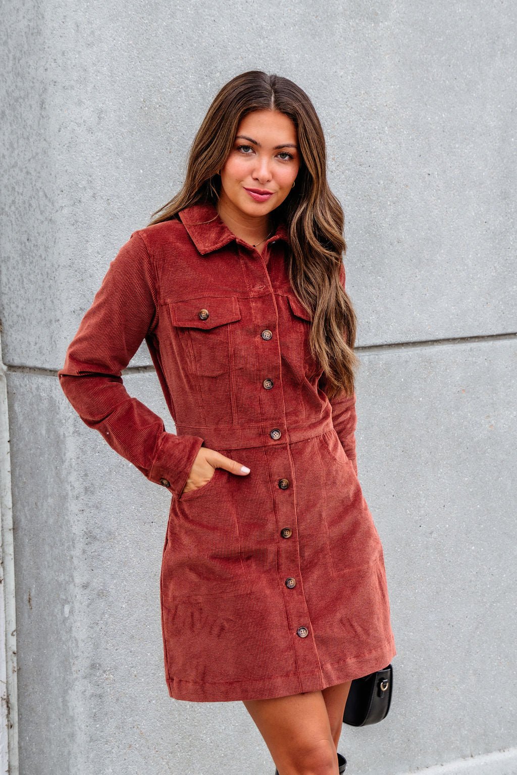 Woman wears the Thread and Supply Rust Corduroy Mini Dress with long sleeves, standing by a gray concrete wall.