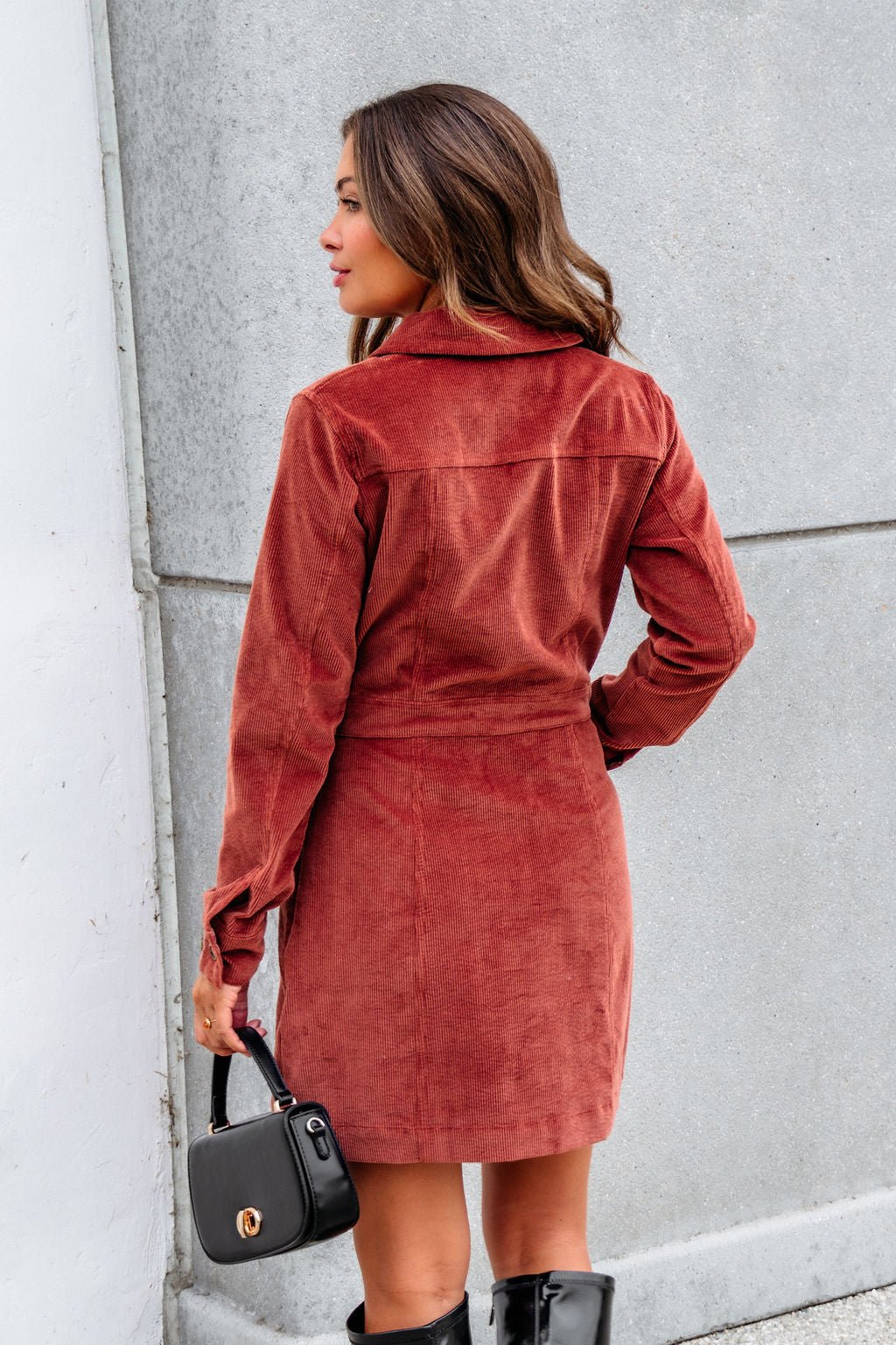 A woman wears the Thread and Supply Rust Corduroy Mini Dress, holding a black handbag and sporting knee-high boots, back to camera.
