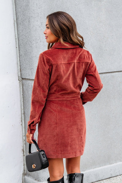 A woman wears the Thread and Supply Rust Corduroy Mini Dress, holding a black handbag and sporting knee-high boots, back to camera.