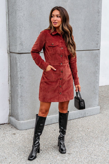 Woman in a Thread and Supply Rust Corduroy Mini Dress and black knee-high boots stands outside, holding a small black handbag.