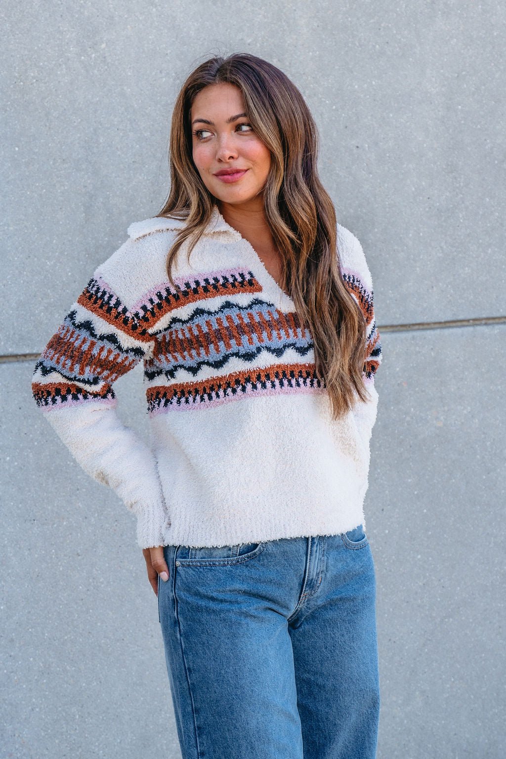 Woman with long brown hair models the Thread and Supply Tan Aztec Bernese Sweater with blue jeans, standing by a gray wall.