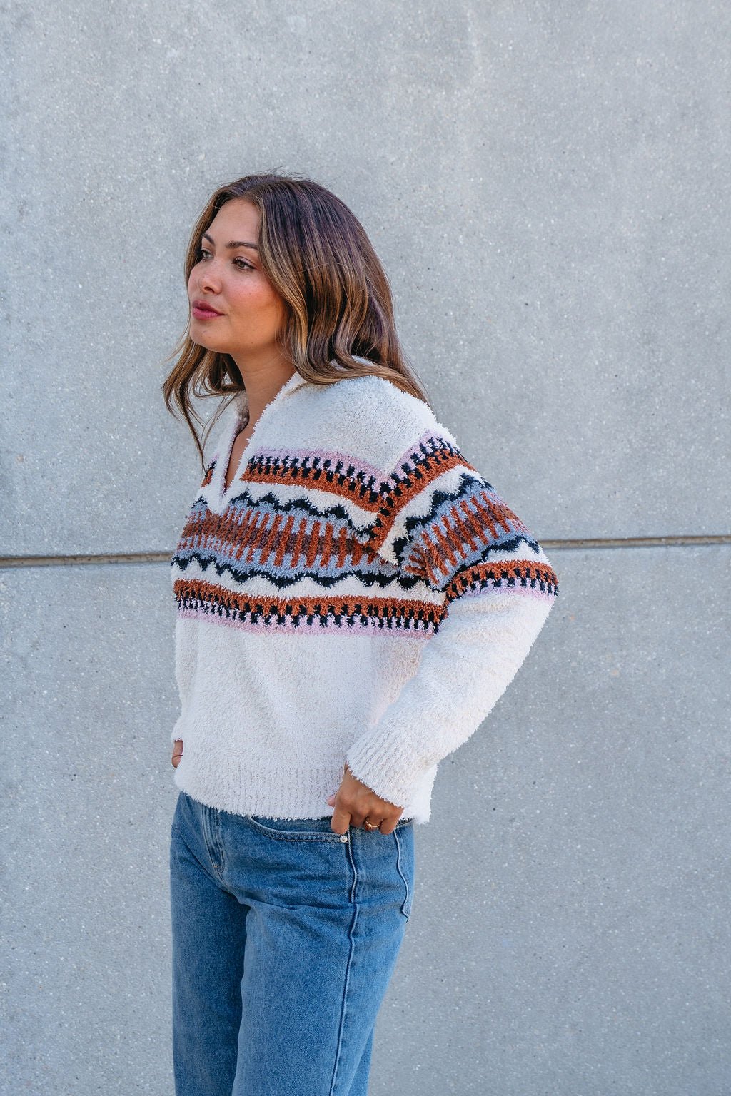 A woman stands sideways against a gray wall, wearing the Thread and Supply Tan Aztec Bernese Sweater and blue jeans.