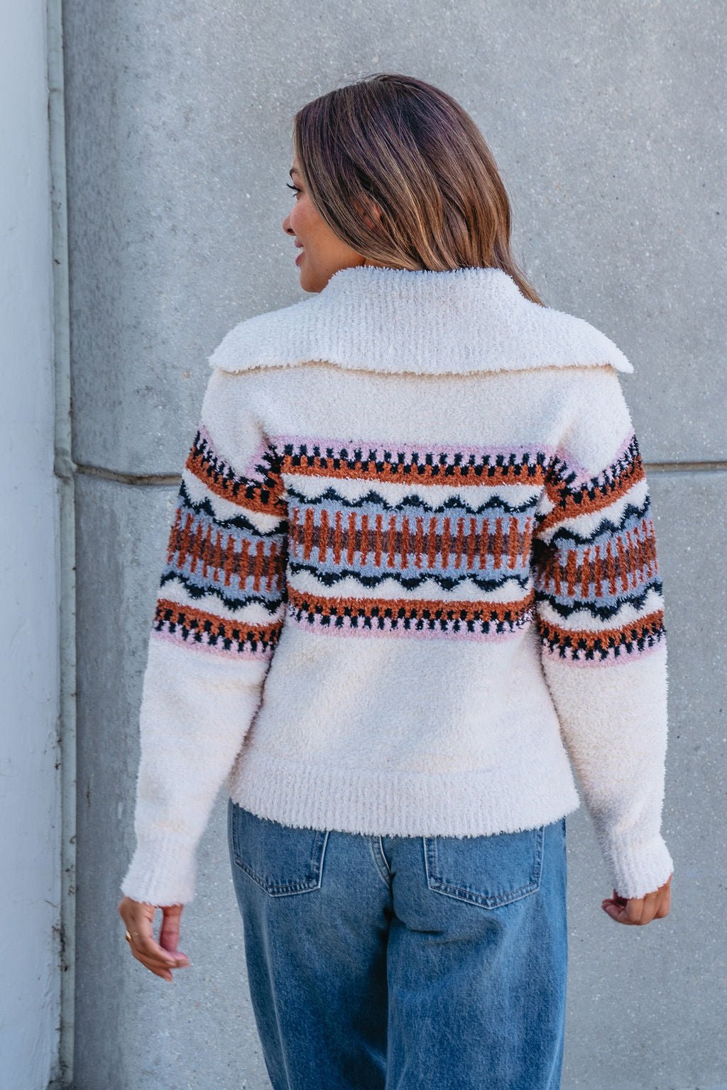 A woman in blue jeans and a Thread and Supply Tan Aztec Bernese Sweater stands with her back to a gray concrete wall.